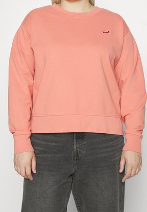 Personne portant un sweat-shirt corail uni à col rond avec un petit logo rouge Levi's sur le haut gauche de la poitrine et un jean noir délavé.