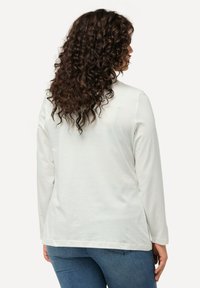 Weißes Langarmoberteil mit glatter Textur, rundem Halsausschnitt und seitlichen Schlitzen, kombiniert mit blauen Jeans. Die Rückansicht zeigt die Passform des Shirts.