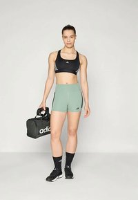 Čierny športový podprsenka s bielymi pruhmi, zelené šortky s bočnými akcentmi, čierne športové topánky a čierny duffel vak s bielym logom.