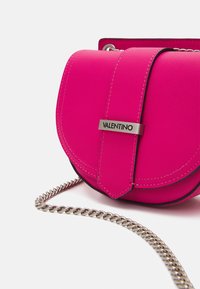 Světle růžová kožená taška přes rameno Valentino ve tvaru sedla s předním p�áskem a stříbrným řetízkovým ramenním popruhem na bílém pozadí.