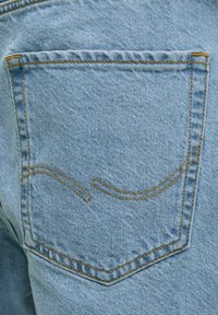 Tylna kieszeń jasnoniebieskich jeansów z pomarańczowym przeszyciem i ozdobnym, zakrzywionym szwem.