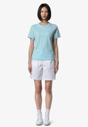 Giovane donna in piedi, indossa una camicia celeste a maniche corte, pantaloncini bianchi fino al ginocchio, calzini bianchi e scarpe da ginnastica bianche.