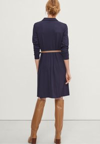 Robe bleu marine avec des manches longues, ceinturée à la taille, dotée d'un col et d'une coupe décontractée. Portée avec des bottes beiges jusqu'aux genoux.