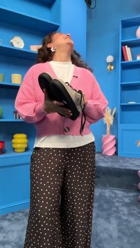 Vrouw in een roze vestje en broek met stippen houdt een paar sneakers vast, glimlacht en kijkt omhoog in een kleurrijke kamer met blauwe planken.