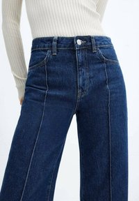 Femme portant un jean taille haute bleu marine à jambes larges et un haut à manches longues crème côtelé, sur fond uni.