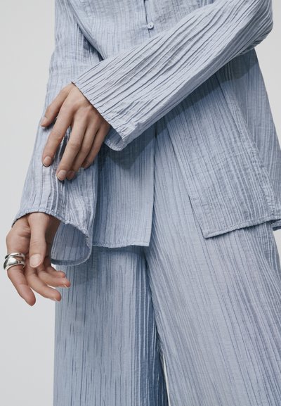 Tecido azul claro, texturizado, com pregas verticais. Inclui uma camisa de botão com colarinho e mangas largas, juntamente com calças descontraídas de corte solto que combinam.