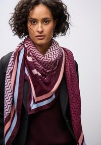 Sciarpa a righe e zigzag nei colori bordeaux e rosa, realizzata in morbido tessuto, drappeggiata attorno al collo su un outfit scuro.