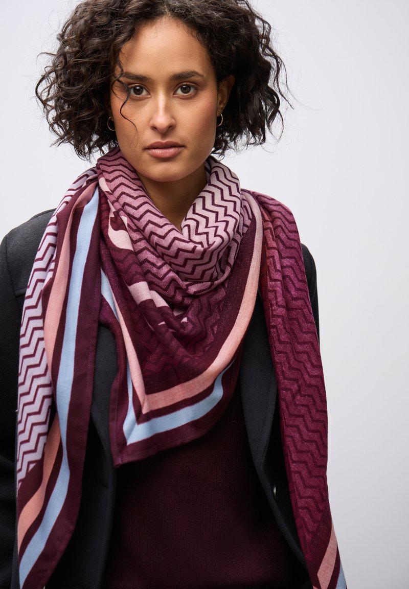 Sciarpa a righe e zigzag nei colori bordeaux e rosa, realizzata in morbido tessuto, drappeggiata attorno al collo su un outfit scuro.