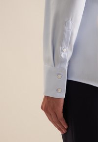 Camicia azzurra chiara in tessuto texturizzato, con polsini arrotondati e tre bottoni bianchi. Mostrata su una mano contro uno sfondo scuro.