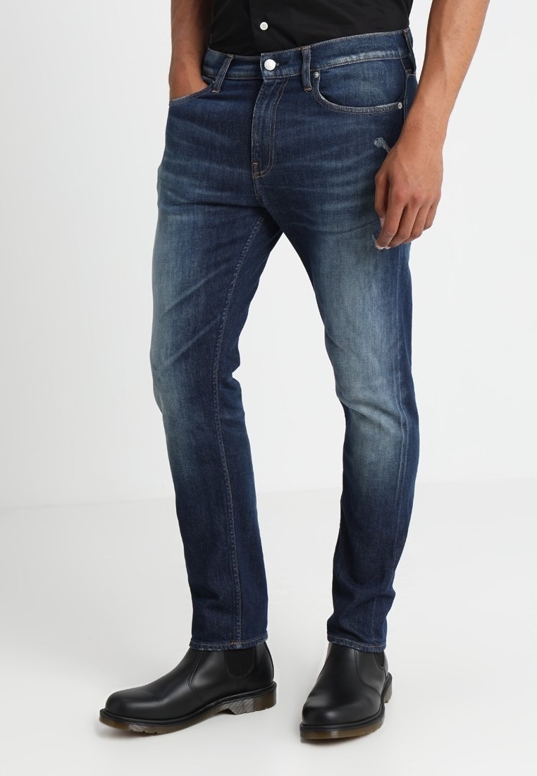 Ciemnoniebieskie dżinsy o dopasowanym kroju, z przetarciami na udach, pięcioma kieszeniami oraz kontrastowymi detalami w szwach. Noszone z czarnymi botkami.