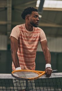 Young man standing behind tennis net holding orange racket, wearing pink striped shirt and white wristbands indoors.