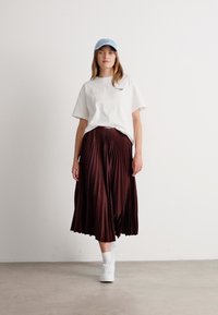 Kvinne med lys blå caps, hvit oversized T-skjorte, plisserte mørkebrune midi-skjørt, hvite sokker og hvite joggesko som går på grått gulv.