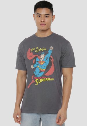 Young man wearing gray t-shirt with colorful Superman graphic and text "This is a job for Superman," standing against plain background.