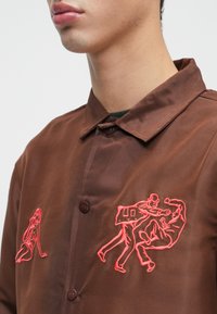 Close-up de uma pessoa a usar uma camisa castanha de botões com bordados vermelhos que representam uma mulher sentada e duas figuras em luta física.