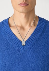 Pull en tricot bleu avec un décolleté en V profond, porté sur un collier en chaîne argenté avec un pendentif rectangulaire à surface décorée.