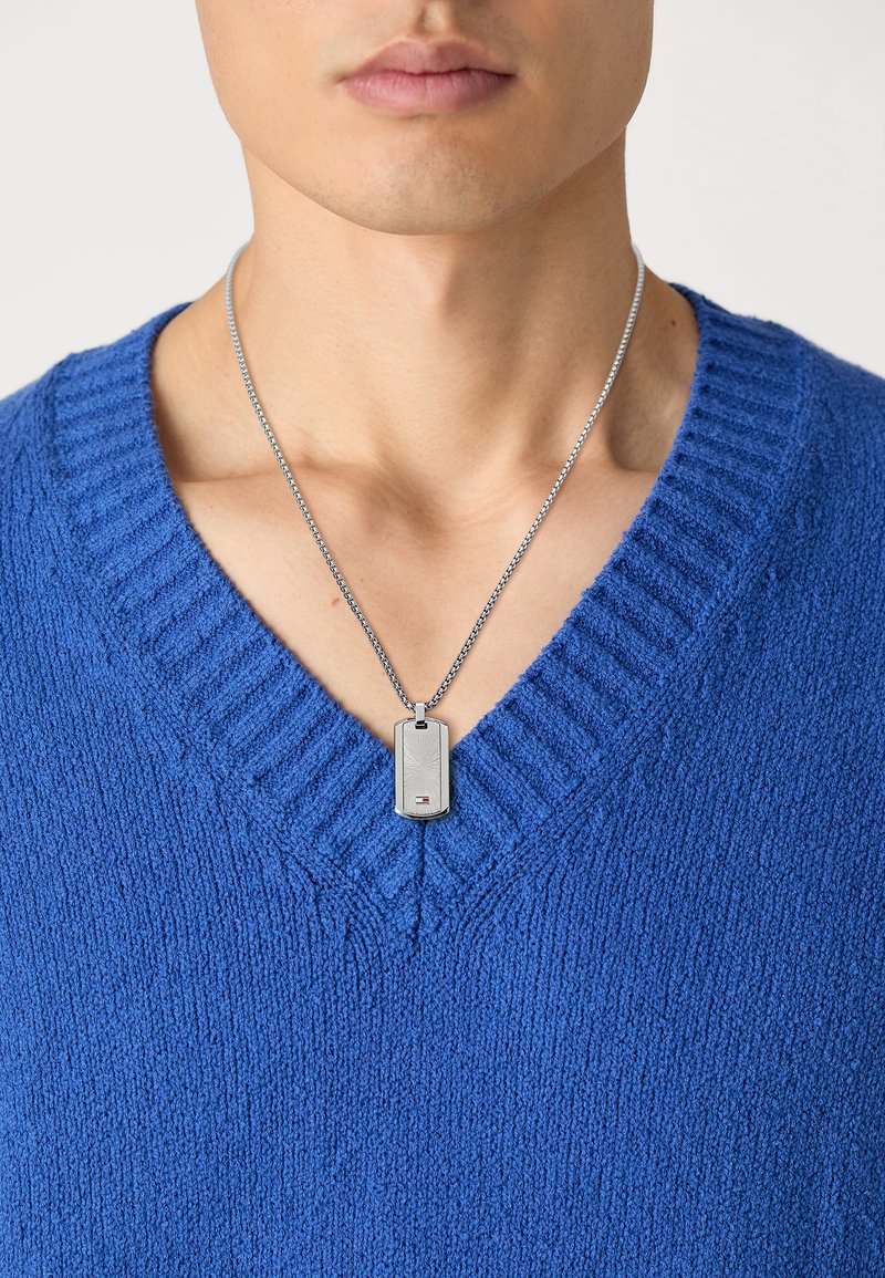 Pull en tricot bleu avec un décolleté en V profond, porté sur un collier en chaîne argenté avec un pendentif rectangulaire à surface décorée.