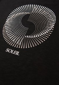 Kreisförmiges Muster aus silbernen Punkten, die ein Spiraldessin über dem Wort "SOLEIL" auf schwarzem Stoff bilden.