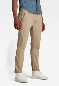 Beige cotton chinos with tapered fit, zip fly, side pockets, and rolled cuffs, paired with white high-top shoes.