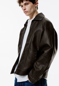 Veste en cuir marron avec un col structuré, fermeture éclair et coupe ajustée. Portée sur une chemise blanche, elle présente un tissu texturé et des coutures visibles.