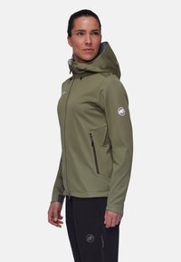 Olivgrüne Softshell-Jacke mit Kapuze, Reißverschlusstaschen und einem Logo auf dem Ärmel. Glatte Textur und figurbetontes Design, kombiniert mit schwarzen Hosen.