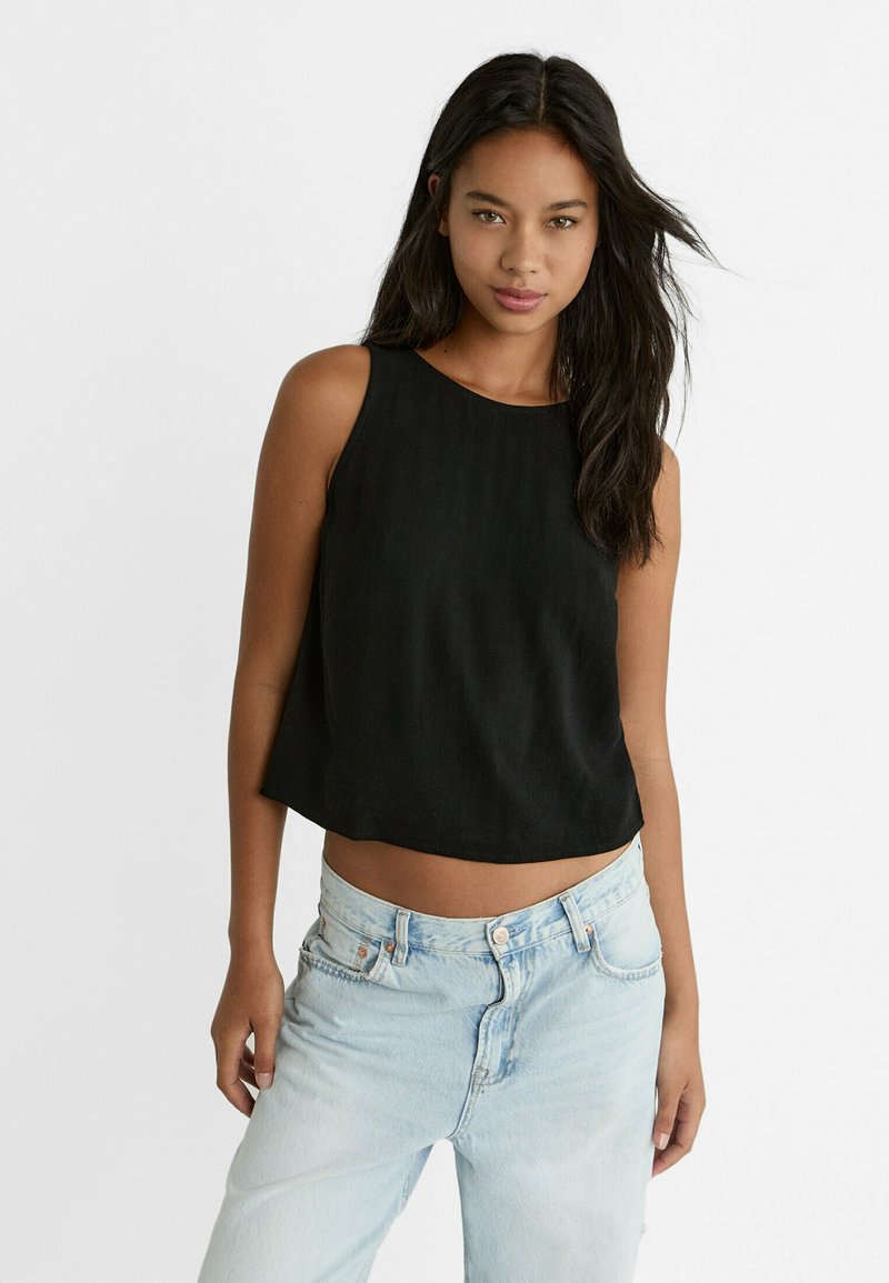 Musta hihaton crop-top, jossa on pyöreä kaula-aukko, valmistettu kevyestä kankaasta. Yhdistetty vaalean sinisiin korkeavyötäröisiin farkkuihin, jotka ovat haalistuneet.