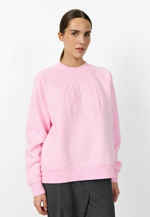 Woman with dark hair tied back wearing a light pink sweatshirt with embossed circular pattern and dark gray pants, standing against white background.