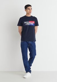 Jūros mėtų spalvos medvilninė marškinė su raudona ir balta "RED BULL RACING" logotipu. Dėvima su tamsiai mėlynais džinsais ir baltais sportbačiais. Paprastas, kasdienis dizainas.