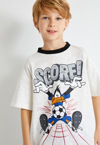 Valkoinen t-paita lyhyillä hihoilla, jossa on sarjakuvamainen ankka hahmo sinisissä ja mustissa jalkapalloasuissa, ja sana "SCORE!" rohkealla harmaalla tekstillä.