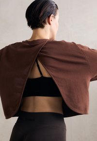 Braunes oversized T-Shirt mit einem geteilten Rückendesign, getragen über einem schwarzen Sport-BH, kombiniert mit dunklen Leggings. Glatte Stoffstruktur.