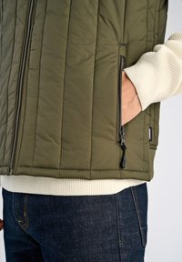 Olivgrüne gesteppte Weste mit horizontalen Nähten, versehen mit einer Reißverschlusstasche und einem cremefarbenen Rippabschluss, getragen über einem hellen Pullover.