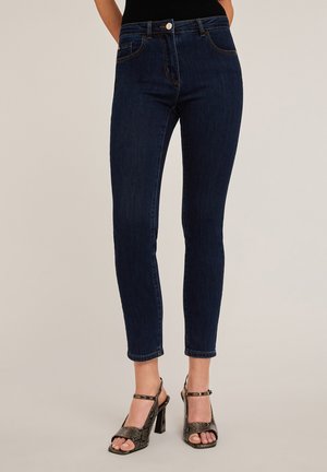 Jean skinny en denim bleu foncé avec cinq poches et une fermeture à bouton à l'avant. Le tissu a une texture lisse et se ressert à la cheville.