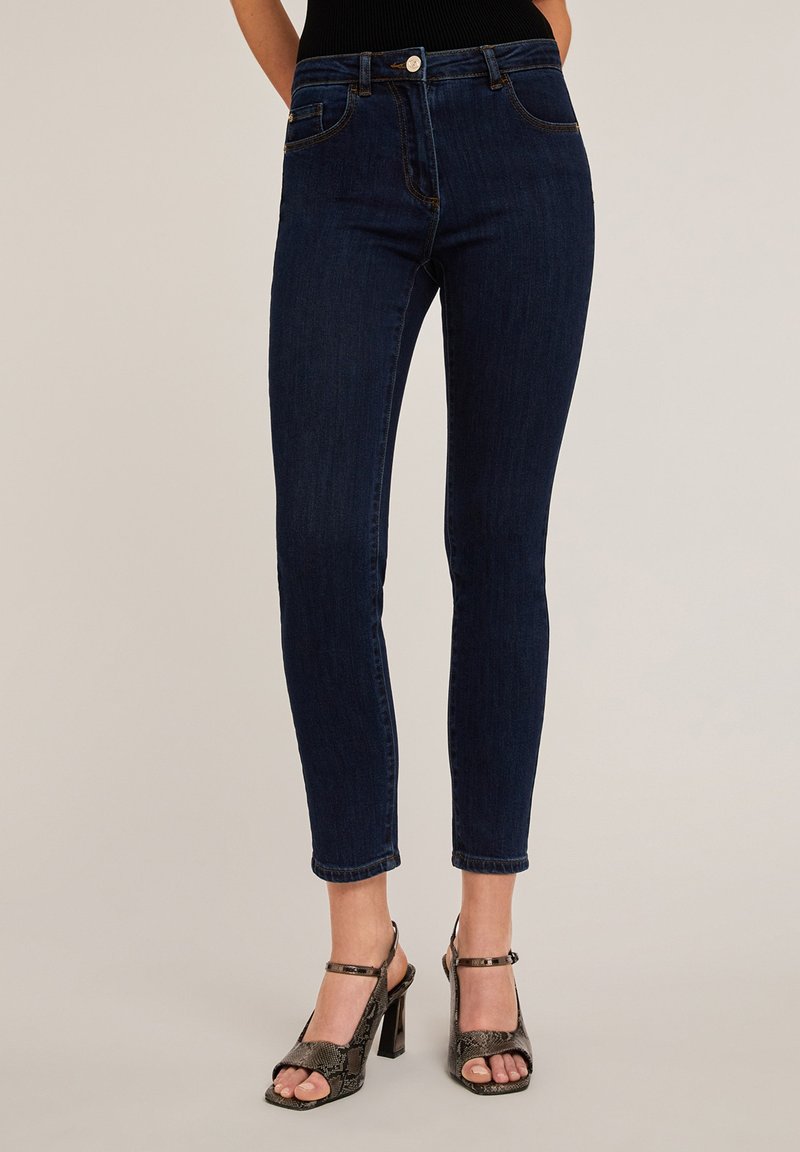 Vaqueros skinnies de mezclilla azul oscuro con cinco bolsillos y cierre frontal de botón. La tela tiene una textura suave y se estrecha en el tobillo.