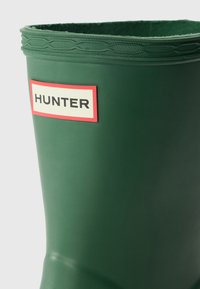 Botas de chuva verdes de borracha com rebordo texturizado e rótulo vermelho com o nome da marca "HUNTER" em preto sobre um fundo claro.