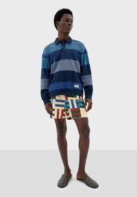 Polo de mangas largas a rayas en azul y marino, combinado con pantalones cortos estampados que presentan rectángulos multicolores sobre un fondo claro.