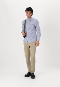 Gestreepte overhemden in blauw en wit, gecombineerd met beige chino's en zwarte schoenen. De persoon houdt een zwarte band over hun schouder.