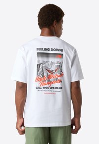 Muž v zelených šortkách, oblečený v bílém tričku s grafikou hory a textem: "Cítíš se sklesle? Vyšší altitude přátelství zavolej 1966-LIFT-ME-UP."