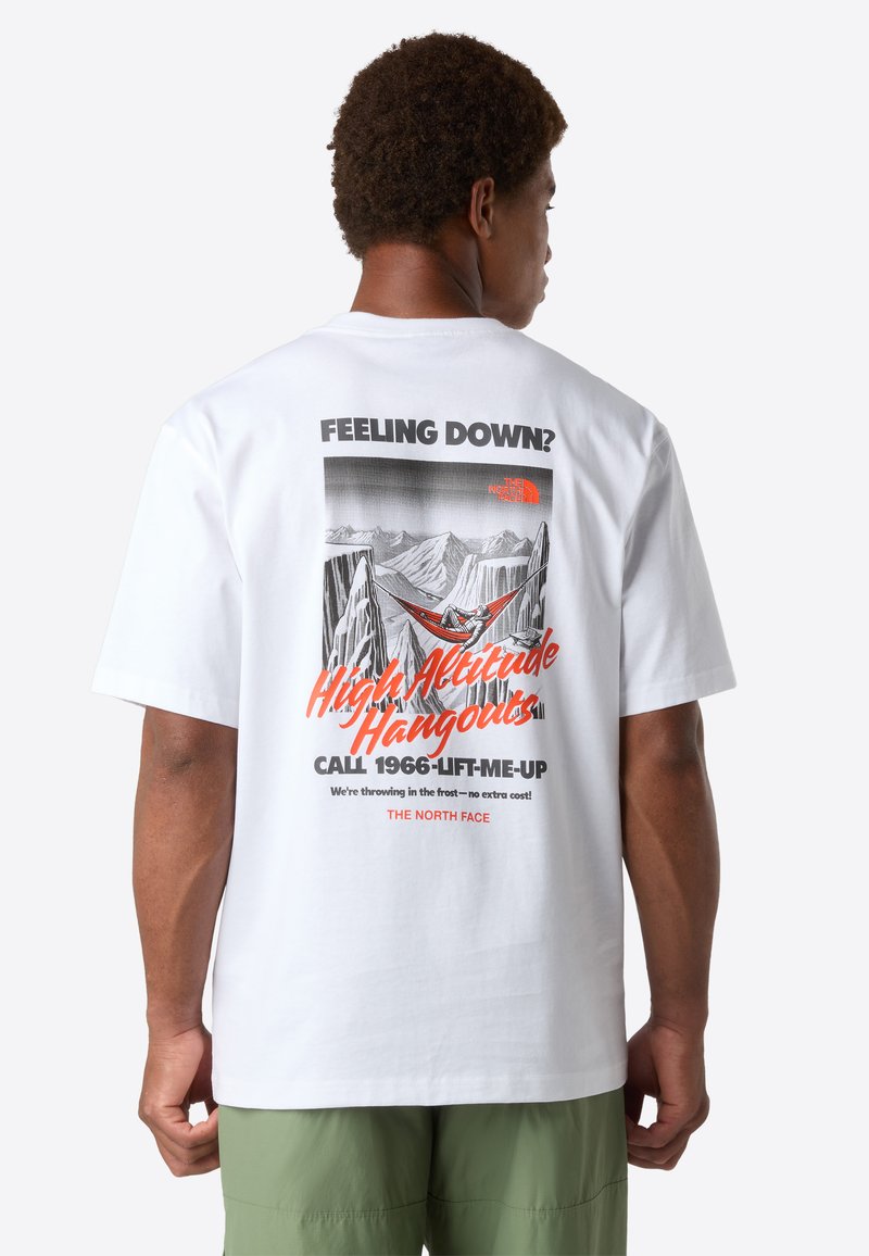 Muž v zelených šortkách, oblečený v bílém tričku s grafikou hory a textem: "Cítíš se sklesle? Vyšší altitude přátelství zavolej 1966-LIFT-ME-UP."