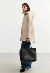 Beige vattentät jacka med huva, frontdragkedja och sidofickor; modell som bär mörkblå vida jeans och håller en svart texturerad tote-väska.