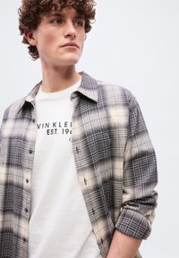 Νεαρός με σγουρά μαλλιά, που φοράει λευκό μπλουζάκι με στάμπα και ανοιχτό γκρι και μπεζ καρό πουκάμισο, σε απλό φόντο.