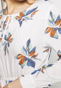 Mujer vistiendo una blusa blanca con estampados florales en naranja, azul y verde, que presenta un escote en forma de llave atado con un pequeño lazo.