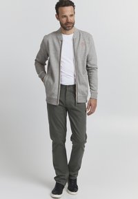 Grau zip-up Jacke mit gerippten Bündchen und Taschen, getragen über einem weißen T-Shirt, kombiniert mit olivgrünen Hosen und schwarzen Turnschuhen.