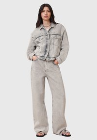Giacca di jeans grigia chiara con orlo a coste e dettagli usurati, abbinata a pantaloni di jeans grigi chiari dalla vestibilità ampia e sandali neri.