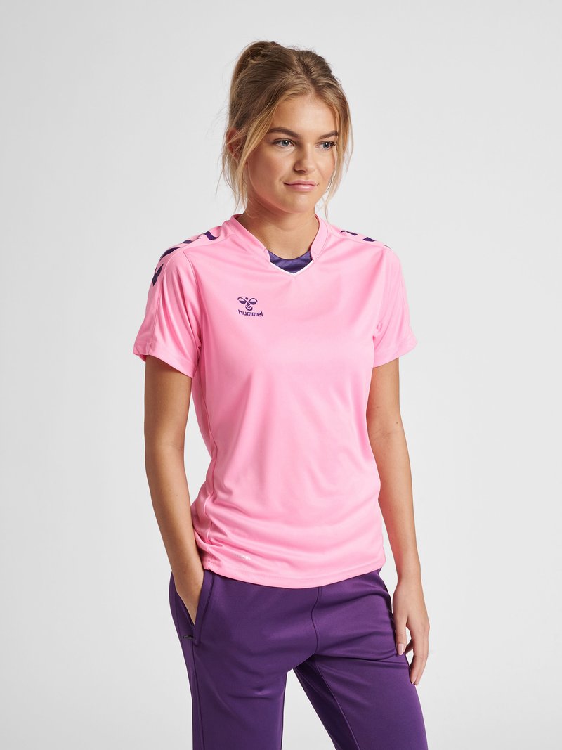 Junge Frau steht mit Händen in den Taschen, trägt ein pinkes Sportshirt und lila Hose vor einfarbigem Hintergrund.