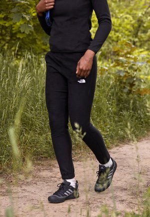 Person iført sorte North Face joggers og sorte Adidas sko går på en grussti omgivet af grønt løv.
