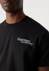 Black cotton t-shirt featuring a round neck and short sleeves; "Espresso Awakening" embroidered in white on the left chest area.