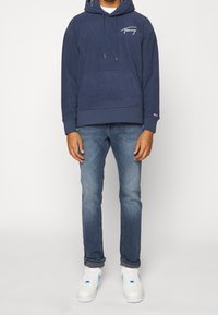 Námořnická mikina s kapucí a přední kapsou, s bílým logem "Tommy", spárovaná s modrými džíny a bílými teniskami s modrými akcenty. Neformální outfit.