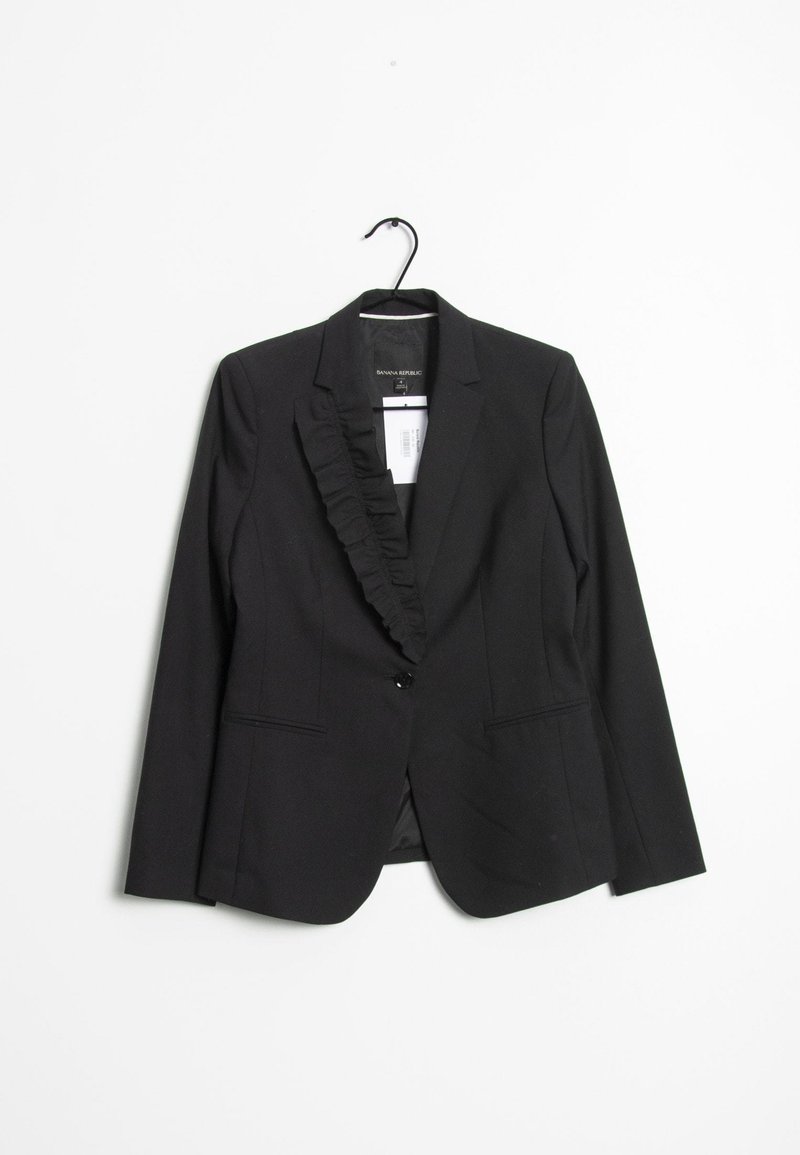 Blazer pour femme noire avec un col à volants, une fermeture à un bouton, deux poches avant et une texture de tissu lisse. Suspendu à un crochet en plastique.
