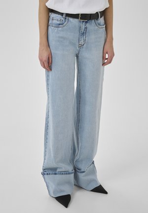 Személy világoskék, széles szárú farmert visel nagyra felhajtott szegéllyel, fekete hegyes orrú cipők fölött, és fehér, beletűrt felsőt.