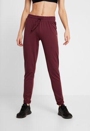 Femme portant un pantalon de jogging bordeaux et des baskets noires, debout jambes écartées sur un fond clair uni.