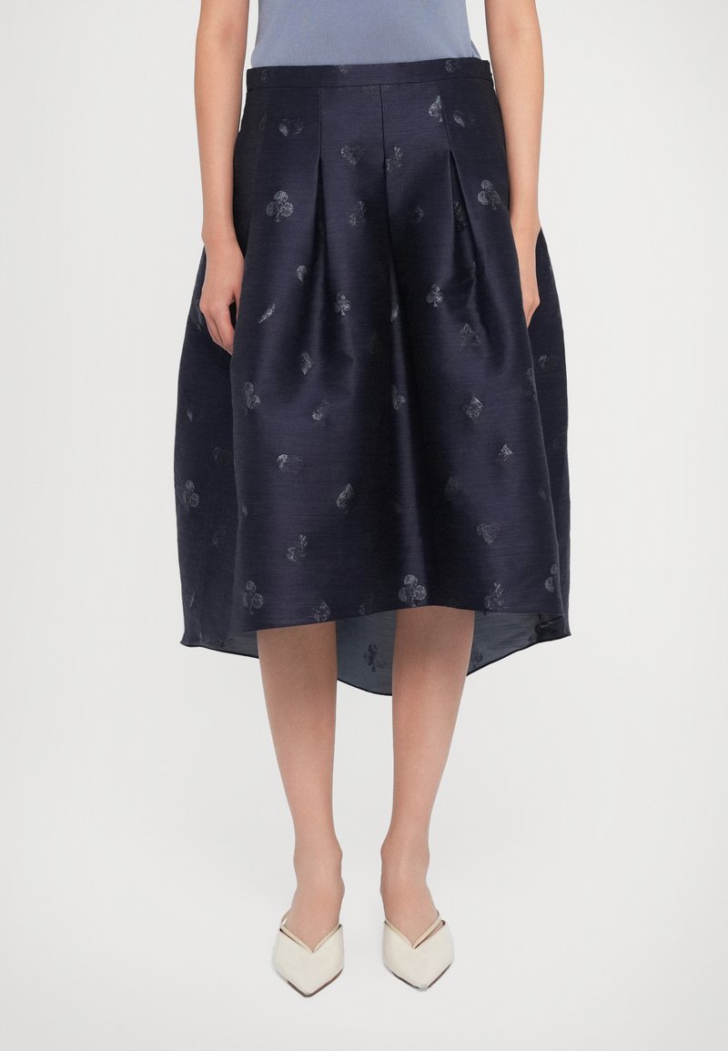 Woman wearing a high-low navy skirt with embossed card suit symbols paired with white pointed-toe shoes, standing against a plain background.